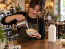 Barista gießt Hafermilch in einen Cappuccino, daneben ein Milchkarton und Haferflocken
