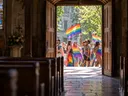 Blick aus einer Kirche durch das offene Portal auf eine fröhliche CSD-Parade mit Regenbogenfahnen
