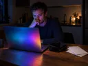 Besorgter Mann sitzt nachts vor einem bunt leuchtenden Laptop, daneben eine leere Geldbörse und Briefe